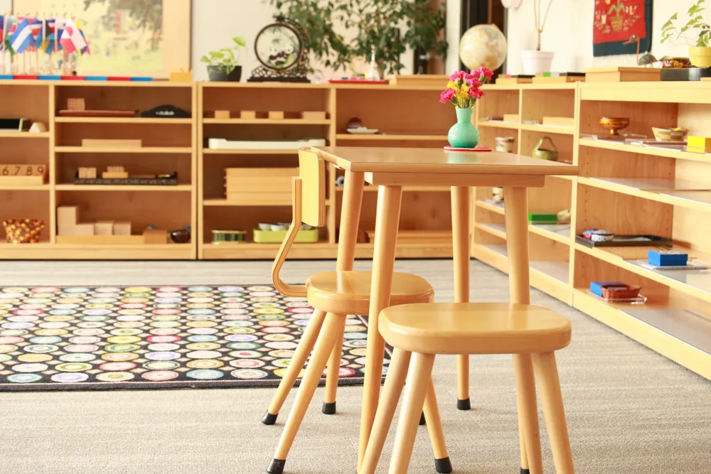 Wooden classroom chairs and shelves of Montessori materials, with a vase of flowers on a low table.