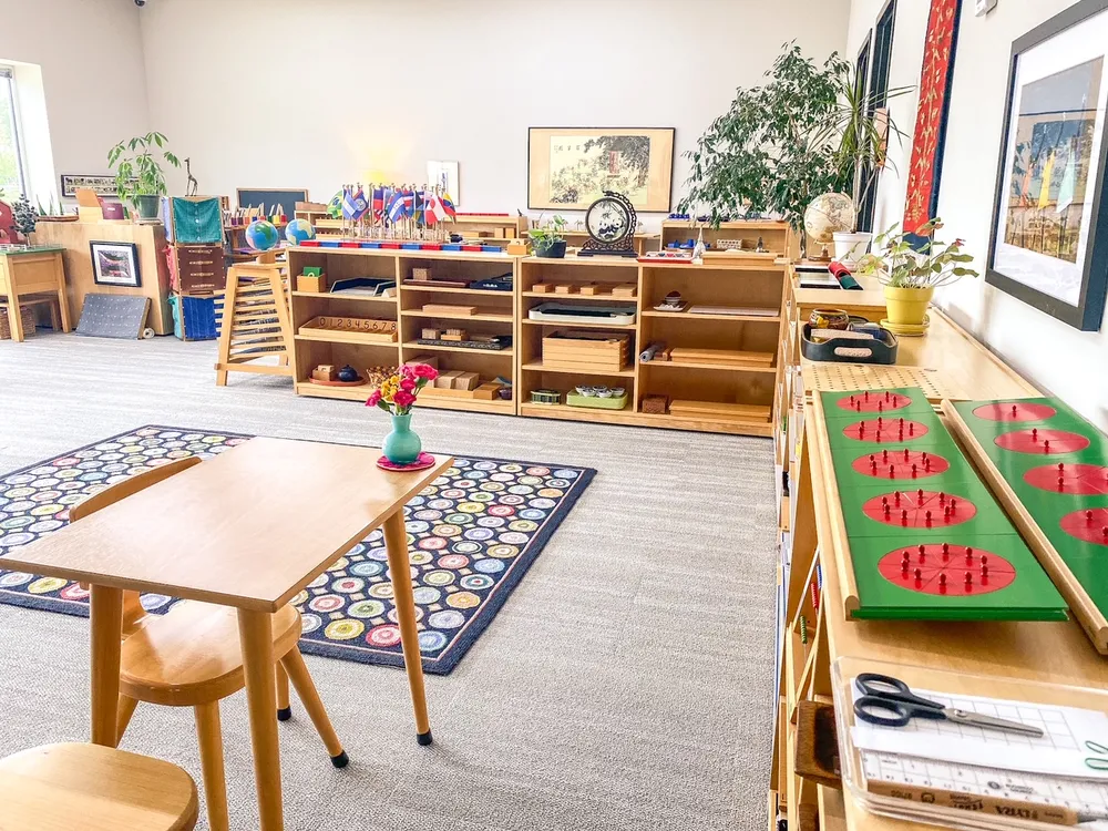 A bright Montessori classroom with low wooden shelves, tables, and carefully arranged learning materials.