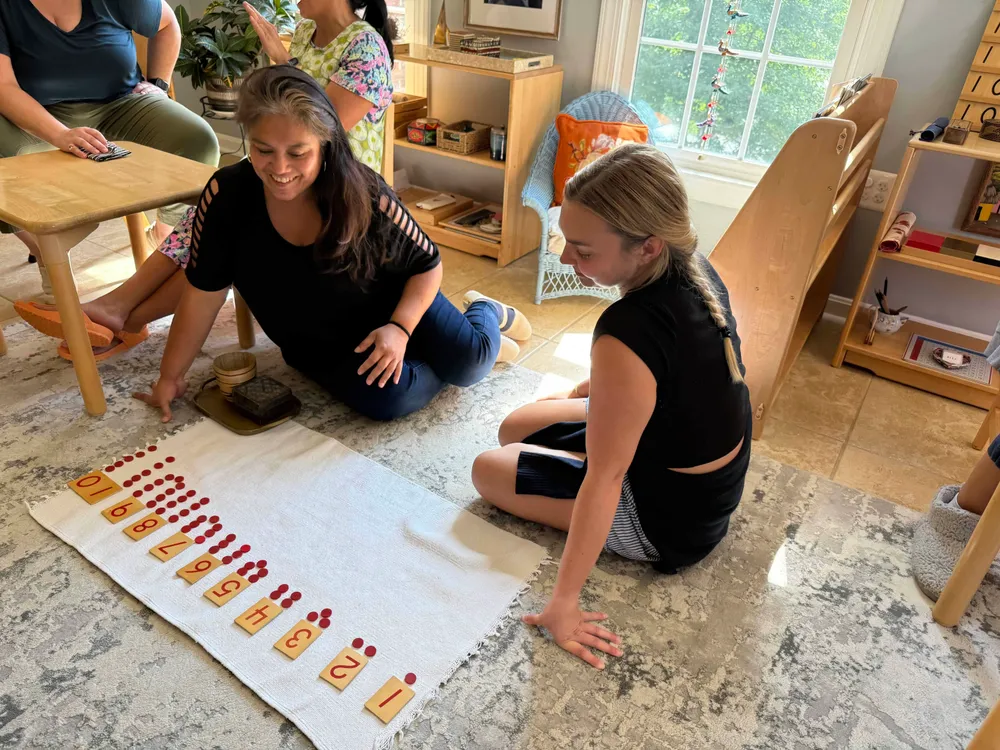 Two adults practicing with numeral cards and red counters on a classroom rug.