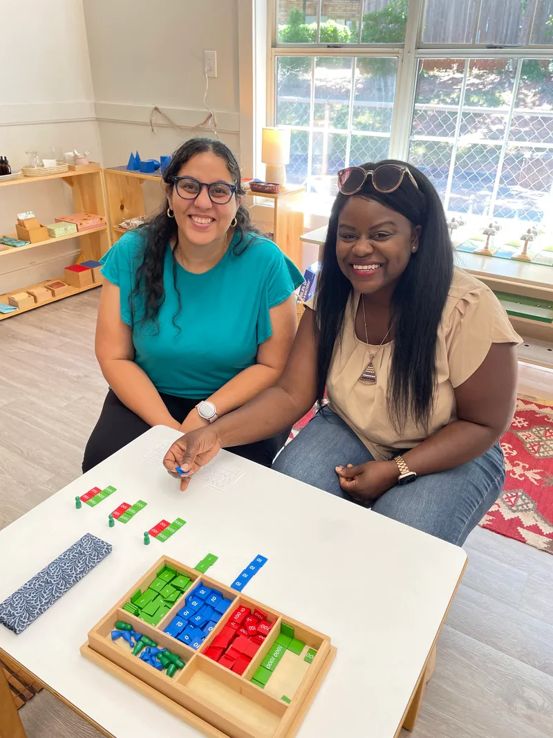 Two smiling adults at a classroom table with the Montessori stamp game and its colored tiles.
